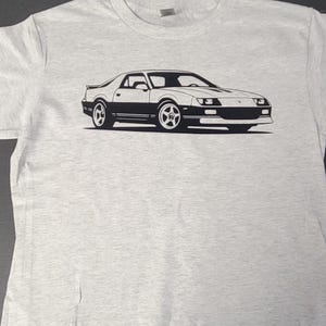 Peut inclure: T-shirt gris clair avec un dessin noir et blanc d'une voiture de sport classique. La voiture est représentée de profil, mettant en valeur son design et ses roues. Le t-shirt est fait d'une matière douce.