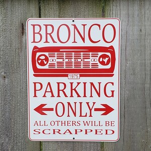 May include: White metal sign with red text that reads "Bronco Parking Only" with a red 1975 Ford Bronco grille graphic and the text "All others will be scrapped".