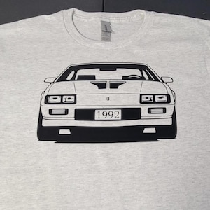 Peut inclure: T-shirt gris clair avec un dessin noir et blanc d'une voiture classique. La vue de face de la voiture est détaillée, avec l'année "1992" affichée sur la plaque d'immatriculation. Le t-shirt a un col rond et des manches courtes.
