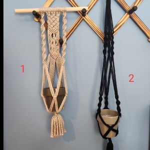 May include: Two macrame plant hangers, one beige and one black, hanging from a wooden rack. The beige hanger has a small terracotta pot with a plant inside. The black hanger has a small terracotta pot with a plant inside. The hangers are labeled "1" and "2".
