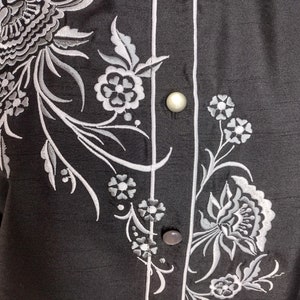 May include: A black button-up shirt with a white floral embroidery design. The shirt has a white trim along the button placket.