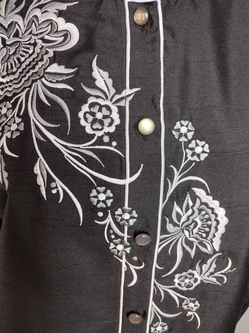 May include: A black button-up shirt with a white floral embroidery design. The shirt has a white trim along the button placket.