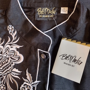 May include: Black button-up shirt with white embroidery of floral designs. The shirt has a mandarin collar and a label that reads "Bob Mackie Wearable Art".