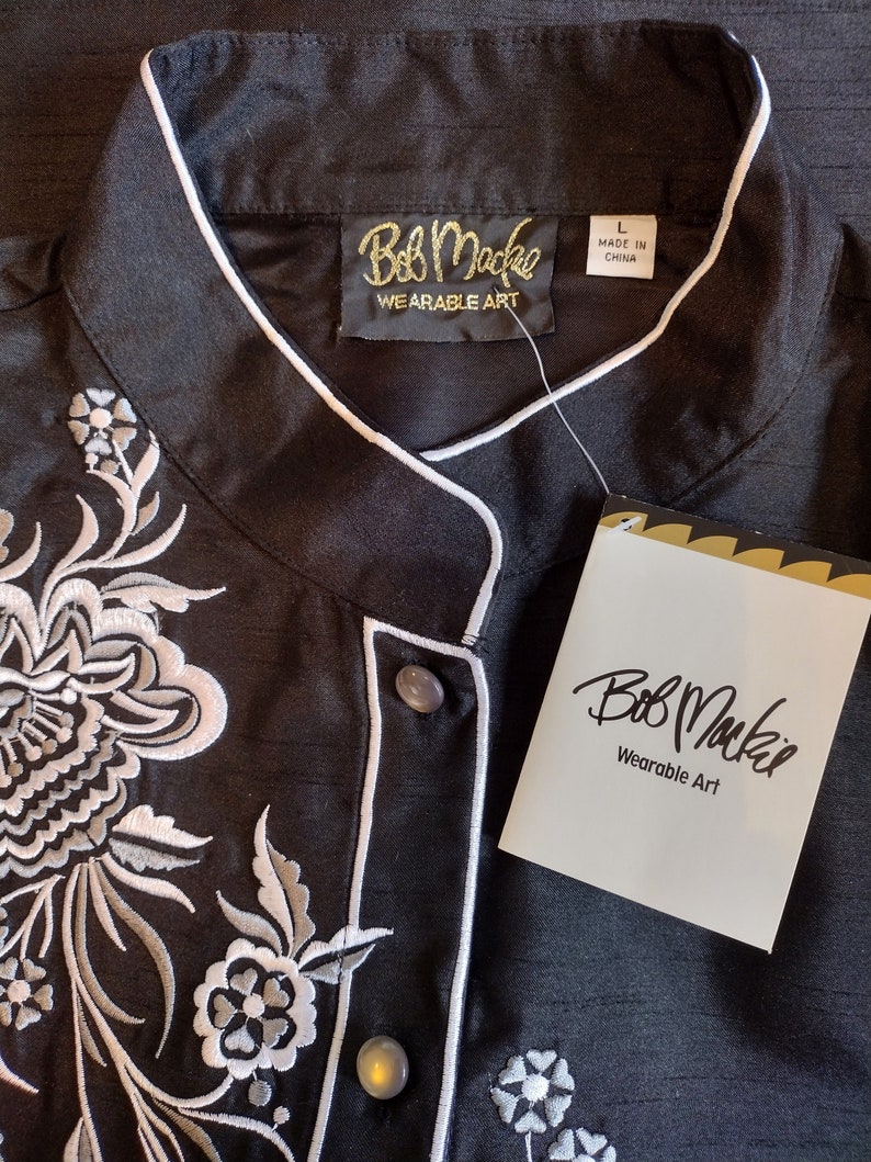 May include: Black button-up shirt with white embroidery of floral designs. The shirt has a mandarin collar and a label that reads "Bob Mackie Wearable Art".