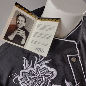 May include: A black embroidered shirt with a white floral design. The shirt has a white collar and a button closure. The text on the tag reads "I've been privileged to create exciting, glamorous fashions for some of the world's most famous and stunning women. A look I've tried to capture in your new Bob Mackie design."  "Each design in my fashion collection is a piece of wearable art, with a whisper of opulence, a hint of extravagance. Each a boldly sophisticated, yet delicately feminine."  "I hope you enjoy your new Bob Mackie design. Wear with your best fashion. Bob Mackie"
