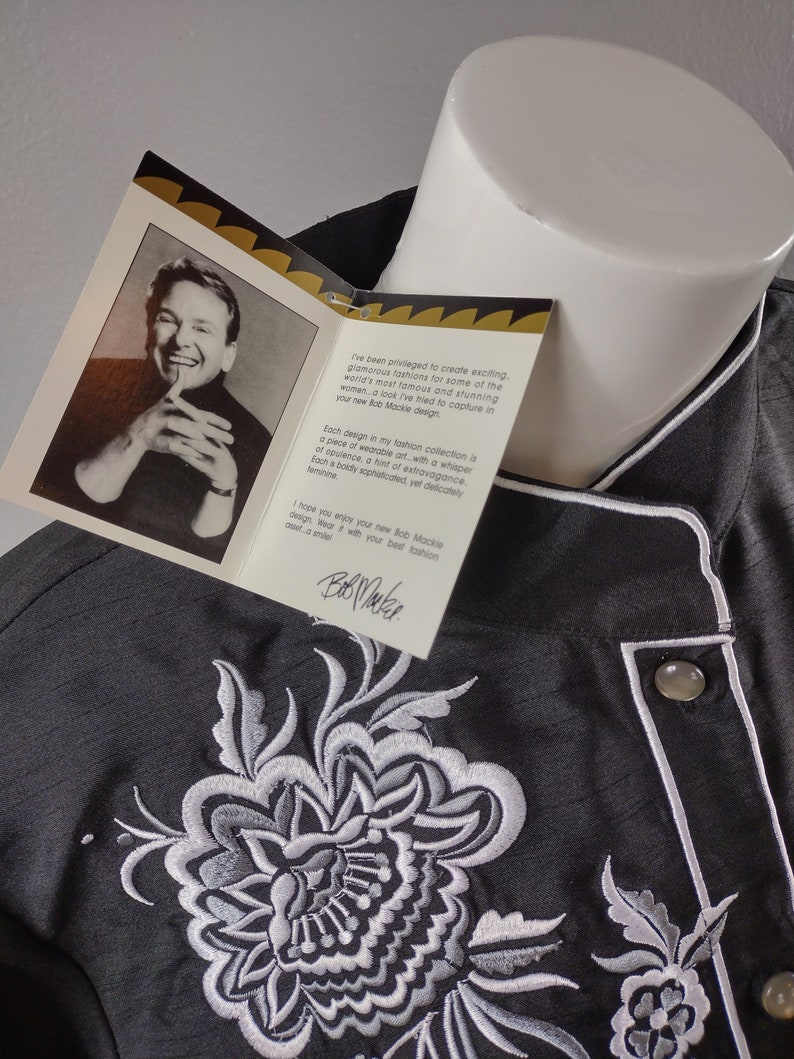 May include: A black embroidered shirt with a white floral design. The shirt has a white collar and a button closure. The text on the tag reads "I've been privileged to create exciting, glamorous fashions for some of the world's most famous and stunning women. A look I've tried to capture in your new Bob Mackie design."  "Each design in my fashion collection is a piece of wearable art, with a whisper of opulence, a hint of extravagance. Each a boldly sophisticated, yet delicately feminine."  "I hope you enjoy your new Bob Mackie design. Wear with your best fashion. Bob Mackie"