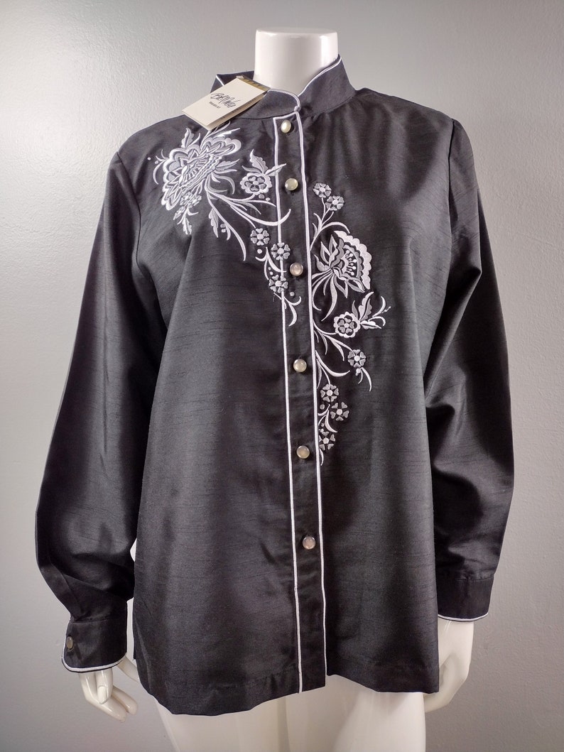 May include: A black long-sleeve button-up shirt with a white floral embroidery design. The shirt has a mandarin collar and a white trim along the front placket and cuffs.