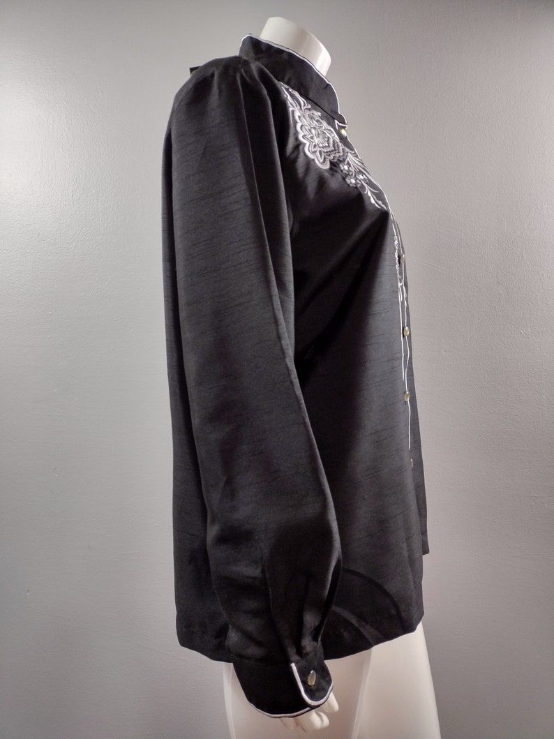 May include: A black long-sleeve shirt with a white embroidered design on the front. The shirt has a mandarin collar and a button-up closure.