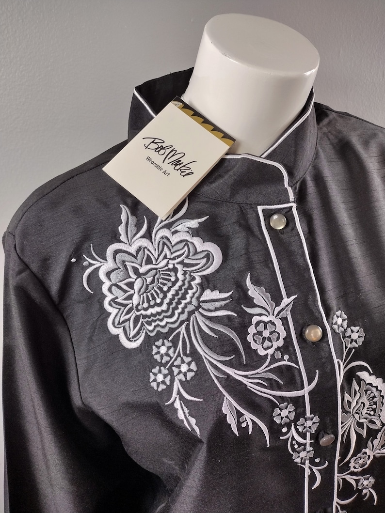 May include: A black long-sleeved shirt with a white embroidered floral design. The shirt has a mandarin collar and a single button closure. The tag on the shirt reads "Bob Mackie Wearable Art."