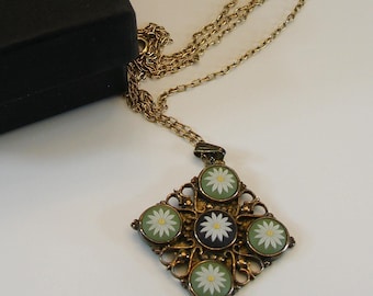 May include: A gold-coloured necklace with a diamond-shaped pendant featuring daisy designs in green and white. The necklace is next to a black box, suggesting it's a gift or for storage. The chain is delicate and the pendant has an ornate frame.