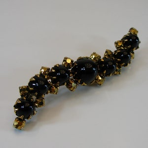 Puede incluir: Broche vintage curvada con una serie de grandes cuentas negras brillantes intercaladas con cristales facetados dorados más pequeños. La broche tiene un engaste de metal dorado, creando un contraste llamativo.