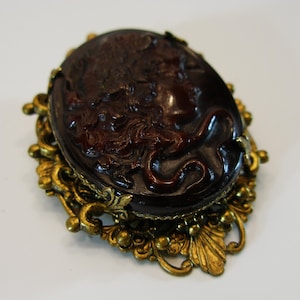 Könnte beinhalten: Ovale, dunkelbraune Kamee-Brosche mit detaillierter Reliefschnitzerei einer klassischen Figur. Die Kamee ist in einem verzierten, goldfarbenen Filigranrahmen gefasst. Ein Accessoire im Vintage-Stil.
