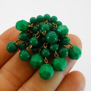 Puede incluir: Broche vintage con un grupo de cuentas verdes de varios tamaños y formas. El broche tiene un respaldo y detalles de cadena de metal dorado, con algunas cuentas facetadas. Las cuentas son de color verde sólido y vibrante.