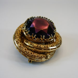 Puede incluir: Un broche dorado con una gran piedra preciosa púrpura iridiscente facetada en el centro. El broche tiene un diseño detallado y ornamentado.