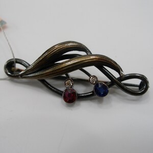 Op de afbeelding: Een donkerkleurige metalen broche met een gebogen, bladvormig ontwerp en een geborstelde bronzen afwerking. Het sieraad is versierd met twee kleine, heldere steentjes en twee grotere, ronde gefacetteerde stenen in donkerrood en donkerblauw.