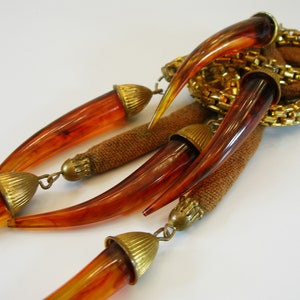 May include: A vintage necklace featuring amber-colored, horn-shaped pendants with brass-toned caps and accents. The design includes a woven brown fabric element and a gold-toned chain detail, creating a unique and eye-catching accessory.