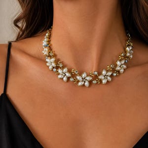 Könnte beinhalten: Eine goldfarbene Halskette mit einer Reihe von miteinander verbundenen Blumenmotiven. Jede Blume besteht aus fünf weißen, marquiseförmigen Kunstperlen mit einem kleinen, klaren, schillernden Kristall in der Mitte, verbunden durch goldfarbene Metallranken.