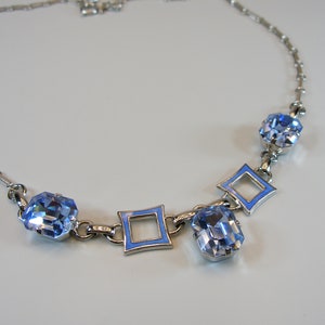 May include: A silver necklace with light blue crystal and enamel details. The necklace has a fine chain with geometric shapes and sparkling blue crystals. The design includes square and oval-shaped blue crystals.