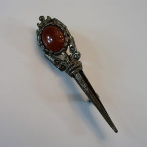 May include: An antique silver-toned kilt pin. It features an oval, reddish-brown stone set in an ornate frame. The pin has a long, pointed design. A decorative accessory, likely for kilts or other garments.