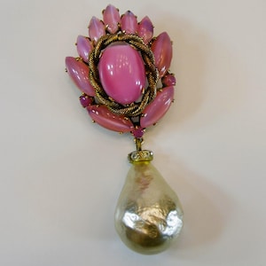 Puede incluir: Broche vintage con una piedra rosa ovalada central, rodeada de piedras en forma de pétalo rosa y morado. Un borde dorado en forma de cuerda rodea la piedra central. Una piedra en forma de pera, iridiscente, blanquecina, cuelga del broche.