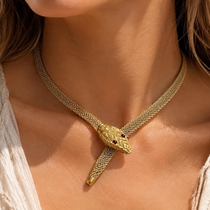 Könnte beinhalten: Eine goldfarbene Schlangenhalskette mit einer flexiblen Netzkettenstruktur. Der Schlangenkopf ist mit texturierten Schuppen und zwei kleinen, dunkelroten Edelsteinaugen versehen. Die Kette liegt um den Hals, wobei der Kopf auf der Kette ruht.