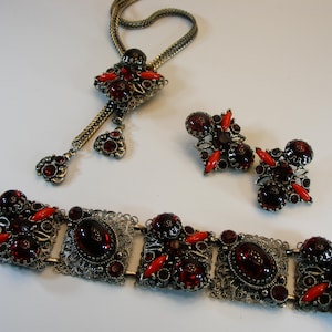 May include: A vintage jewelry set featuring a necklace, earrings, and bracelet. The set has a silver-tone metal base with ornate filigree details. The jewelry is adorned with oval and round dark red stones and bright red accents.