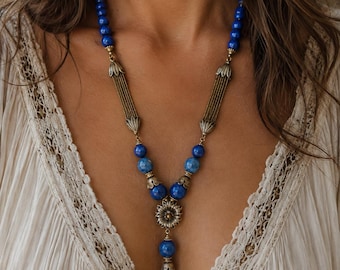 Puede incluir: Un collar con cuentas azules redondas y detalles en metal color latón antiguo. El diseño incluye dos barras metálicas verticales estriadas, tapas decorativas en forma de flor y un colgante central con forma de flor y una cuenta colgante.