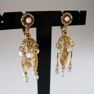 May include: A pair of gold-toned earrings with a floral design and white beads. The earrings have a filigree bell shape with dangling white beads and gold leaf-shaped accents. The earrings are displayed on a black stand.
