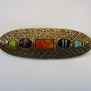 May include: An oval gold-coloured brooch with a detailed woven design. It features five stones in various shapes and colours: green, purple, orange, black and white, and blue. A vintage accessory, suitable for a coat or scarf.