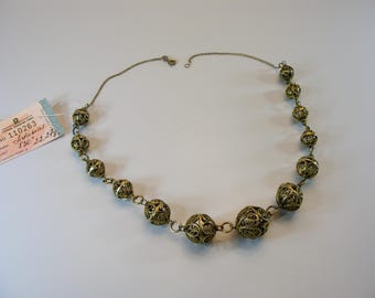 May include: A vintage gold-toned filigree necklace with a series of ornate, spherical beads connected by delicate chain links. The necklace is displayed on a white surface, with a price tag attached. The tag has text in a foreign language.