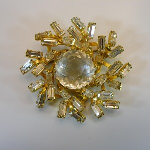 May include: A gold-toned brooch with a central, large, clear, faceted crystal surrounded by smaller, rectangular, clear crystals. The brooch has a sunburst design, with the crystals radiating outwards.
