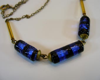 Op de afbeelding: Een vintage ketting met blauwe en zwarte cilindrische kralen met een dambordpatroon. De kralen zijn verbonden door goudkleurige metalen staven en een delicate ketting. De ketting wordt getoond op een wit oppervlak.