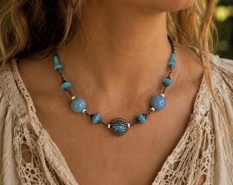 May include: A necklace featuring a series of light blue glass beads and small white spacer beads strung on a dark metal chain. The centerpiece is a round, textured dark metal bead with a light blue inlay, flanked by blue beads and metal caps.