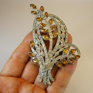 Könnte beinhalten: Silberfarbene Brosche in einem Bouquet-Design, verziert mit klaren und bernsteinfarbenen Strasssteinen. Die Brosche hat ein detailliertes, durchbrochenes Design mit einer bandgebundenen Basis, wodurch ein funkelndes, elegantes Accessoire entsteht. Die Brosche ist etwa 7.6 cm lang.