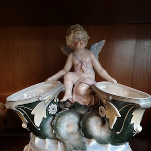 Puede incluir: Centro de mesa de cerámica con un querubín sentado sobre dos cuencos abiertos. Los cuencos están sostenidos por una base con diseños de delfines y olas, y la base está decorada con motivos florales. La paleta de colores incluye blanco, verde, azul y rosa.