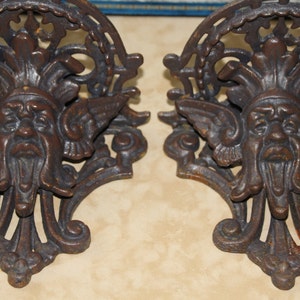 May include: Two antique, cast iron wall sconces with a dark brown, aged finish. Each sconce features a detailed face with wings and ornate scrollwork. The design is reminiscent of classical or gothic architectural elements.
