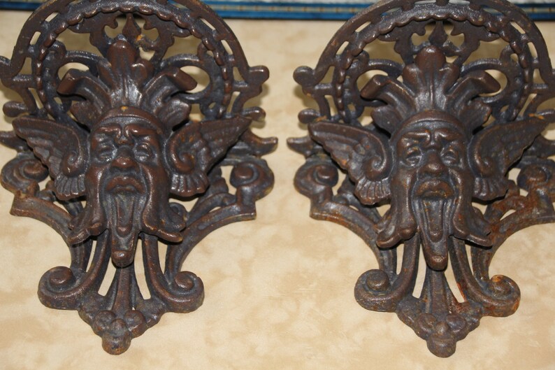 May include: Two antique, cast iron wall sconces with a dark brown, aged finish. Each sconce features a detailed face with wings and ornate scrollwork. The design is reminiscent of classical or gothic architectural elements.