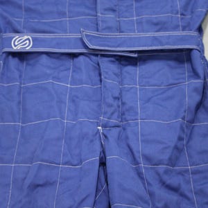 May include: Close-up of a blue racing suit with white stitching. The suit has a belt with a logo and a grid-like pattern. The fabric appears to be a durable material, suitable for motorsports.