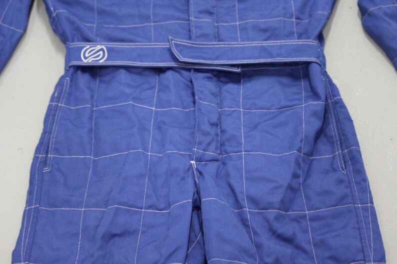 May include: Close-up of a blue racing suit with white stitching. The suit has a belt with a logo and a grid-like pattern. The fabric appears to be a durable material, suitable for motorsports.