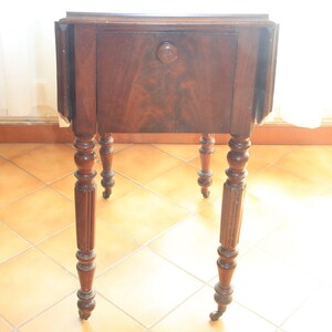 Puede incluir: Una mesa de madera marr&oacute;n oscuro con un solo caj&oacute;n y patas torneadas. La mesa tiene una tapa abatible a cada lado y est&aacute; sobre un suelo de baldosas.