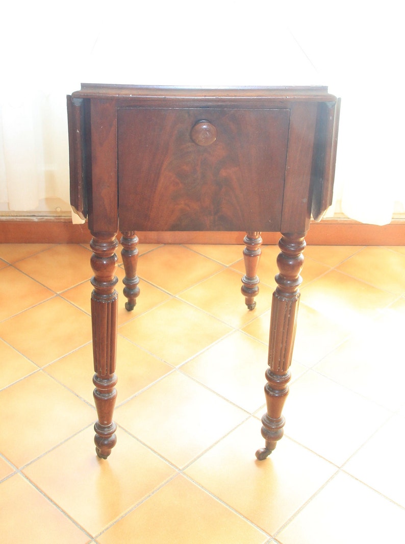 Puede incluir: Una mesa de madera marr&oacute;n oscuro con un solo caj&oacute;n y patas torneadas. La mesa tiene una tapa abatible a cada lado y est&aacute; sobre un suelo de baldosas.