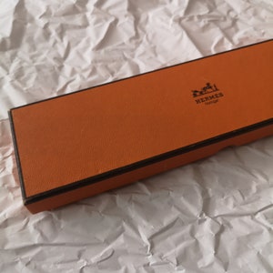 Könnte beinhalten: Eine orangefarbene Schachtel mit braunem Rand. Auf der Schachtel befindet sich das Hermes-Logo und der Schriftzug "Horloger".