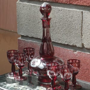 Könnte beinhalten: Ein Set aus rubinrotem Glas, darunter eine Karaffe und sechs Gläser. Die Karaffe hat einen runden Boden und einen langen Hals, der von einem dekorativen Verschluss gekrönt wird. Die Gläser und die Karaffe haben ein weiß geätztes Design.