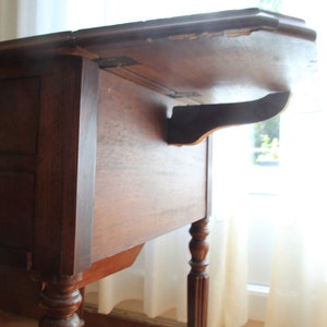 Puede incluir: Una mesa de madera marr&oacute;n oscuro con una tapa abatible y un caj&oacute;n. La mesa tiene patas torneadas y est&aacute; en buen estado.