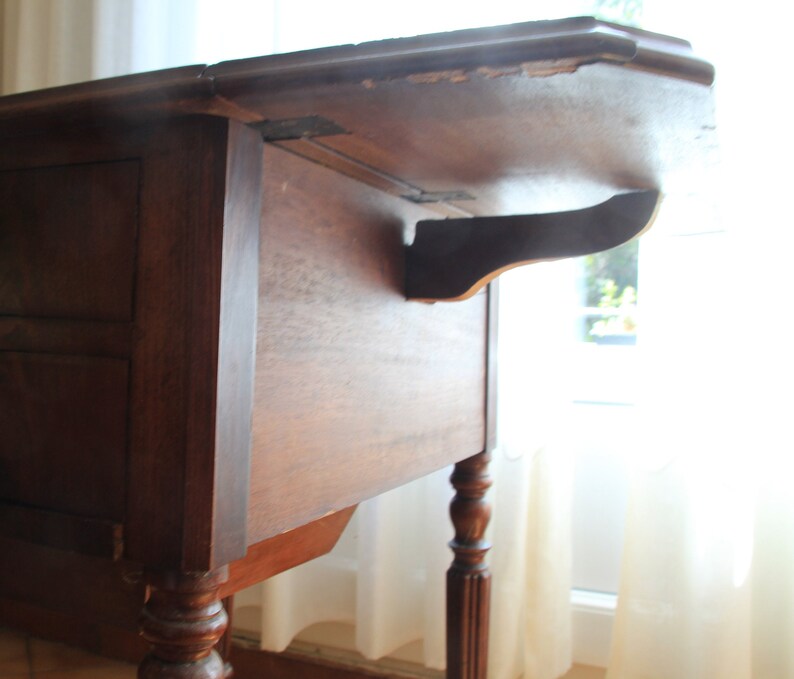 Puede incluir: Una mesa de madera marr&oacute;n oscuro con una tapa abatible y un caj&oacute;n. La mesa tiene patas torneadas y est&aacute; en buen estado.