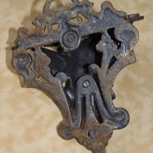 May include: Ornate, cast iron door knocker with a dark gray finish and visible rust. The knocker features a decorative, floral design with a central, stylized shape. The metal shows signs of age and wear, adding to its vintage appeal.