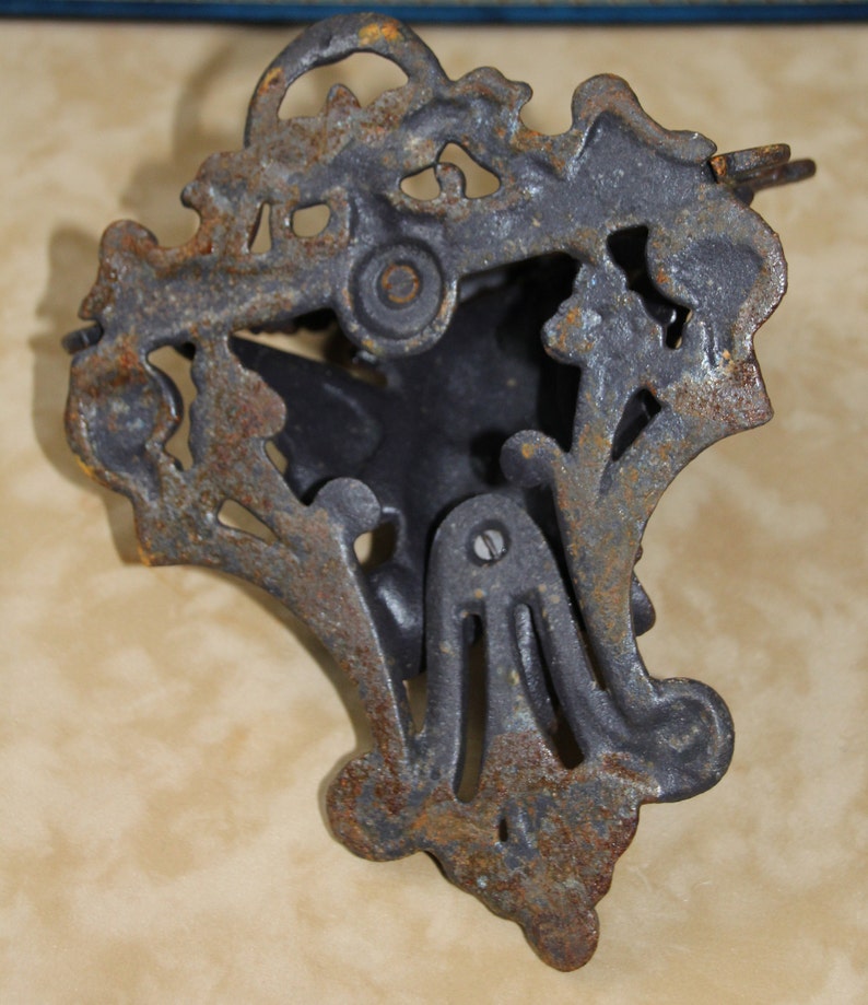 May include: Ornate, cast iron door knocker with a dark gray finish and visible rust. The knocker features a decorative, floral design with a central, stylized shape. The metal shows signs of age and wear, adding to its vintage appeal.
