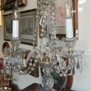 May include: A clear crystal chandelier with two candle-shaped light bulbs. The chandelier features crystal pendants and a decorative base. The background includes framed artwork.