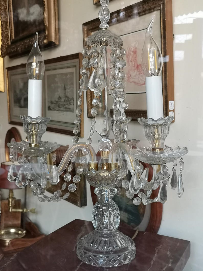 May include: A clear crystal chandelier with two candle-shaped light bulbs. The chandelier features crystal pendants and a decorative base. The background includes framed artwork.