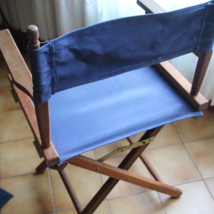May include: A wooden director's chair with a blue canvas seat and back. The chair is folded and sitting on a tiled floor.